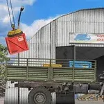 Gri bir kamyon, bir hangarın önünde duruyor, yukarıda vinçle kaldırılan kırmızı bir konteyner var